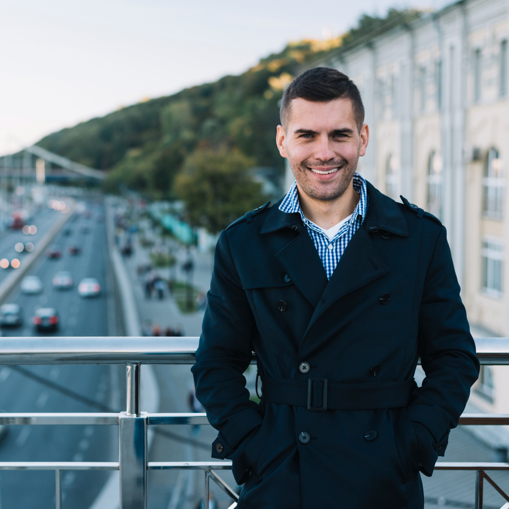 Person wearing a dark coat and checkered shirt, smiling on bridge.