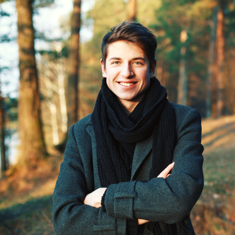 Person in a dark coat and scarf smiling in a forest setting.