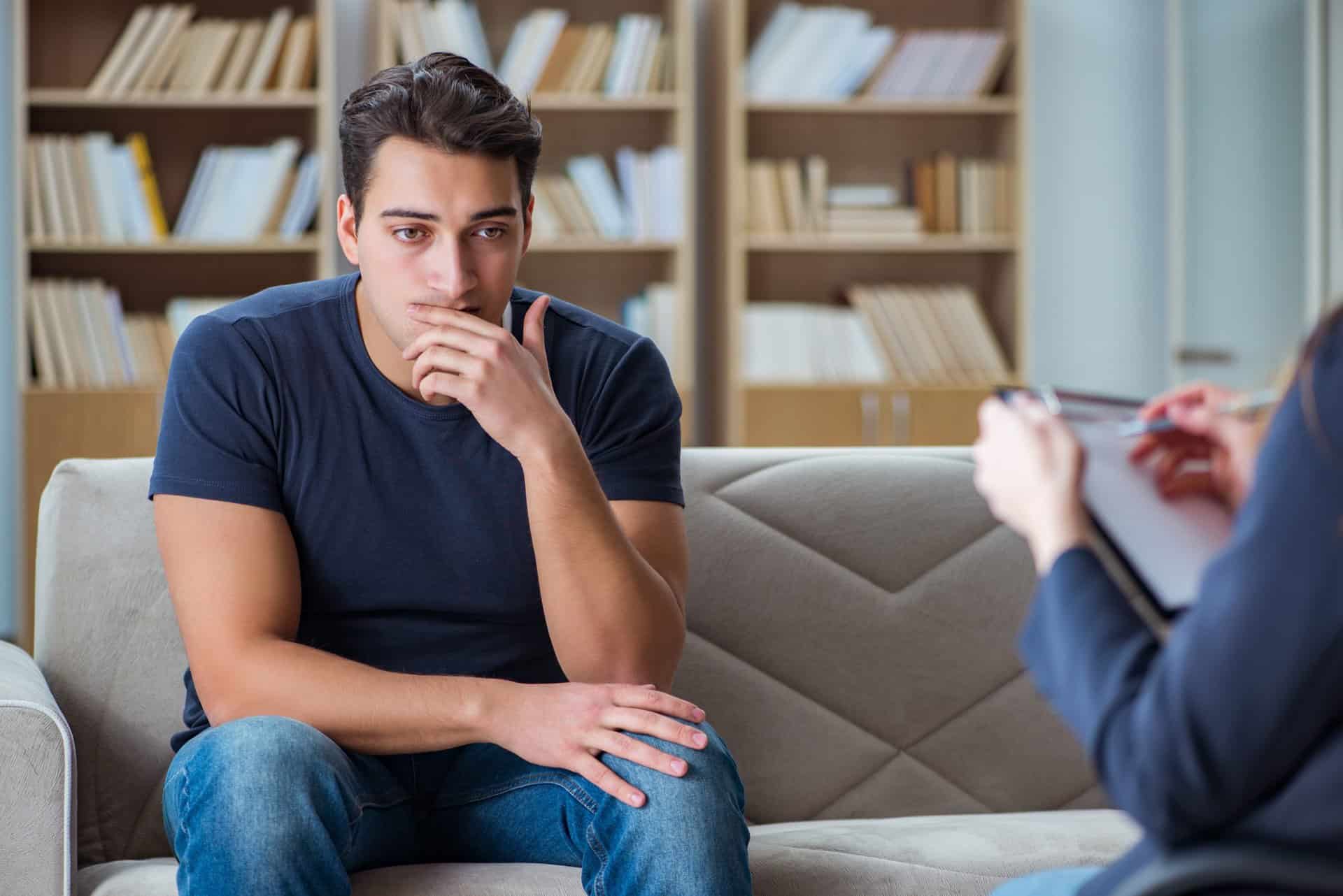 Person wearing a navy t-shirt and jeans sitting on couch, hand to mouth, in thoughtful pose.