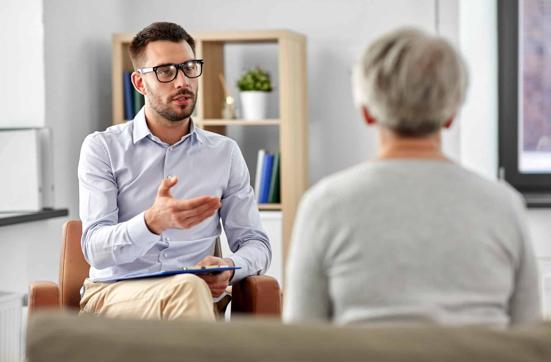 Person in glasses with blue shirt holding clipboard, speaking to another person with grey hair.