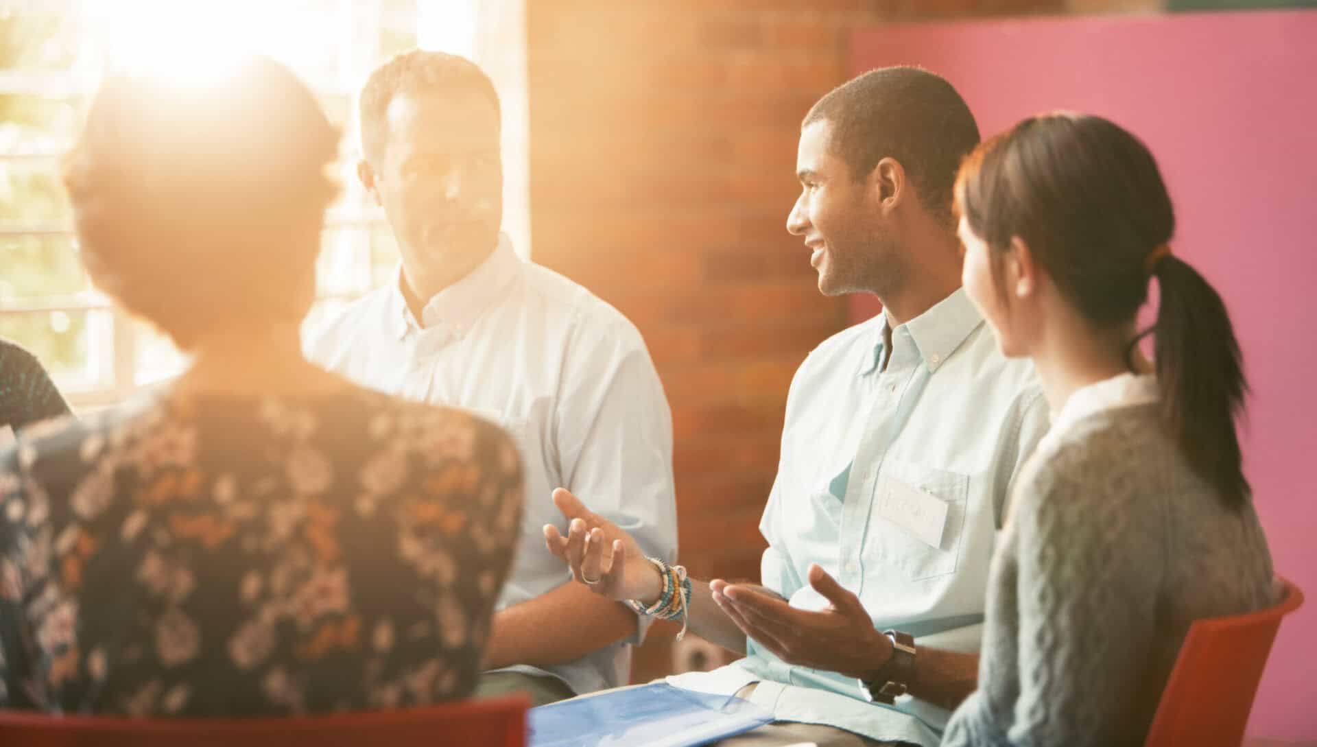 Group of people in a discussion, one person gesturing with hand, others listening, in a sunlit room.