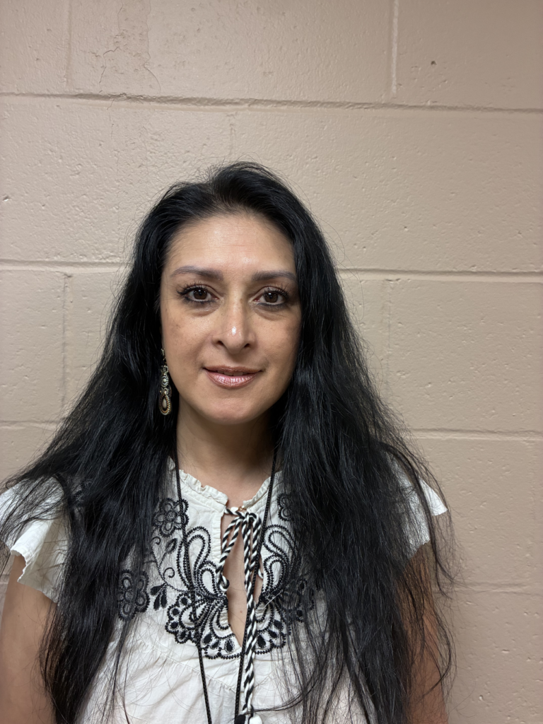 Person wearing a white blouse with black embroidery, long hair, standing against a beige wall.