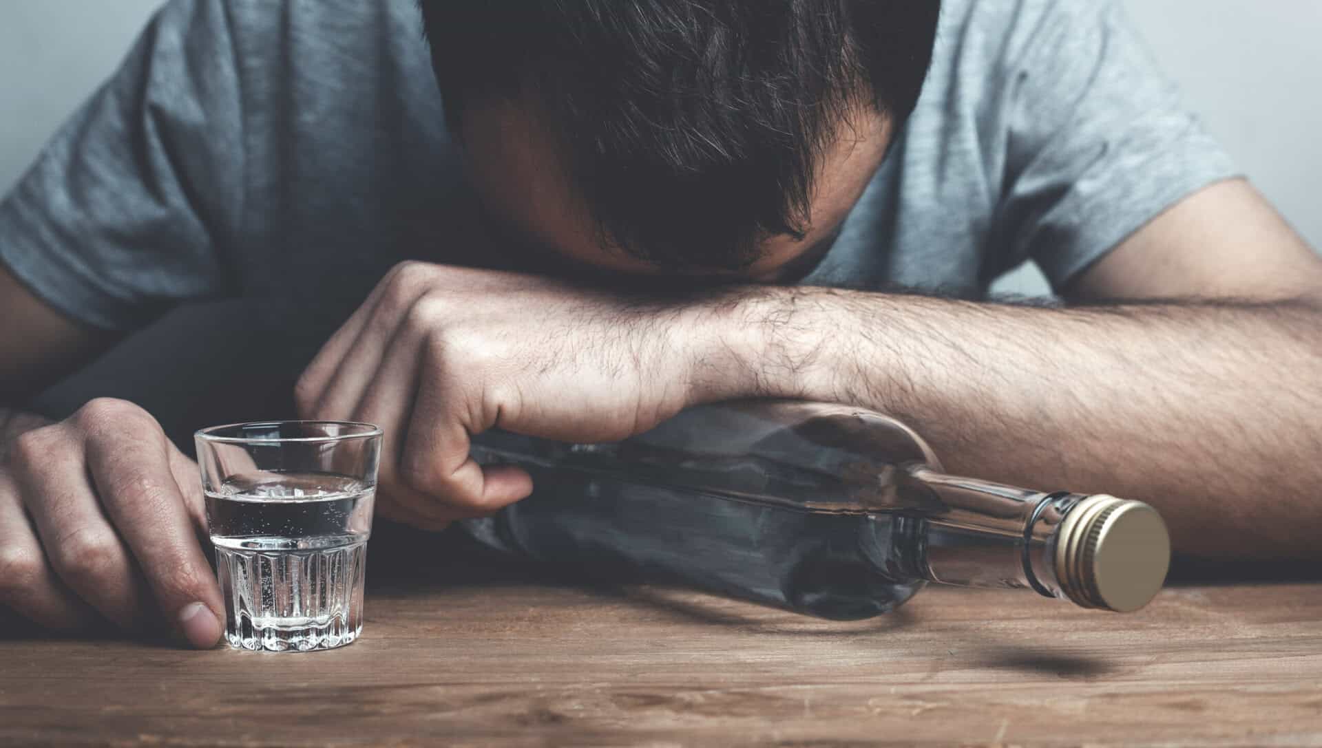 Person wearing a grey shirt with head on arm, holding a bottle next to a shot glass.
