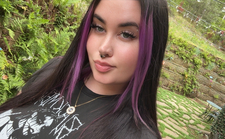 Person with long dark hair and purple streaks wearing a graphic t-shirt and necklace, standing outdoors.