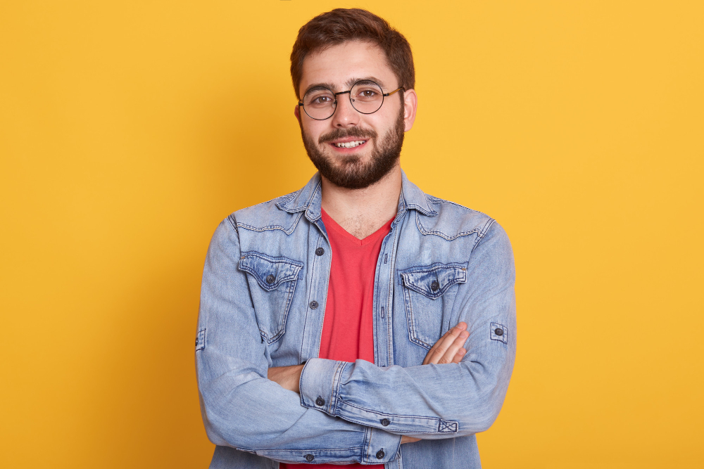 Person wearing glasses and a denim shirt with arms crossed against yellow background.