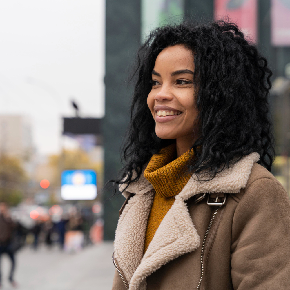 Person wearing a brown coat with a shearling collar and a mustard sweater, smiling outdoors.