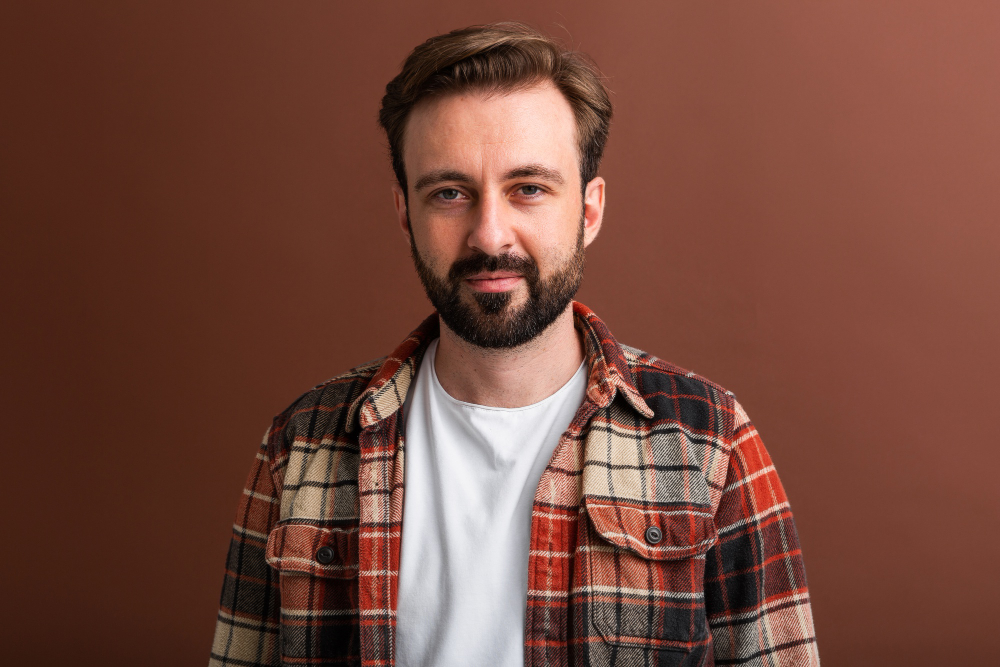Person wearing a plaid shirt and white t-shirt, with a beard and neutral expression.