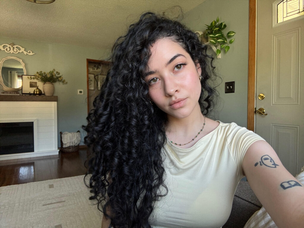 ```plaintext
Person with curly hair in a cream shirt sitting indoors
```